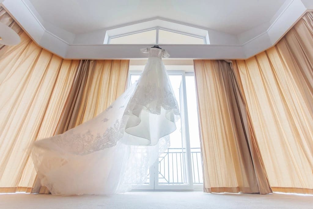 Vestido de renda e tule, de saia ampla e cauda curta, pendurado diante de uma janela entre cortinas cor de areia