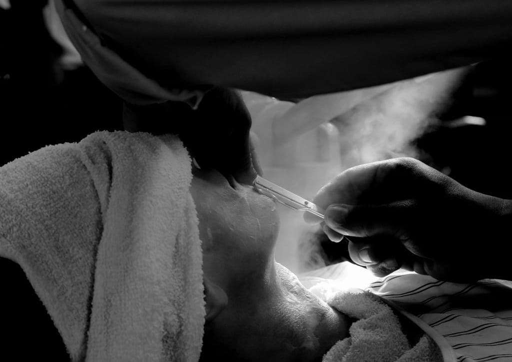 Toalha quente sobre o rosto durante a barba, com vapor e a mão do barbeiro com a navalha